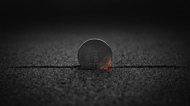 A coin with the Bitcoin mark stands upright in dark sand, resting exactly on a single straight line drawn across the sand, with darkness above.